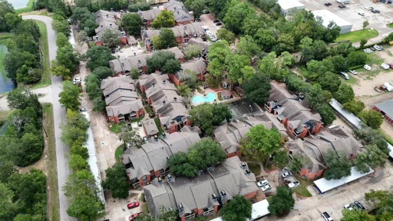 Aerial view of an apartment complex with grey roofs, trees, and a central swimming pool.